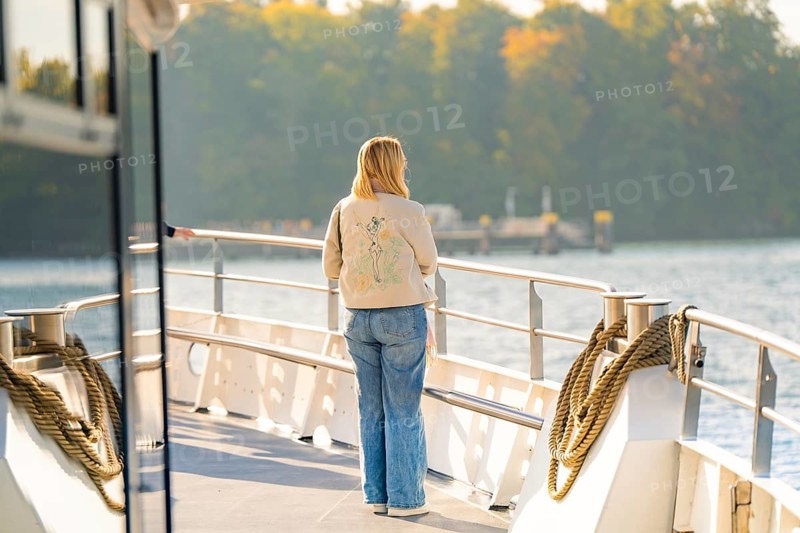 Woman on the deck of a boat looking at water and autumn trees in the distance, boat trip, Lake Constance, Germany