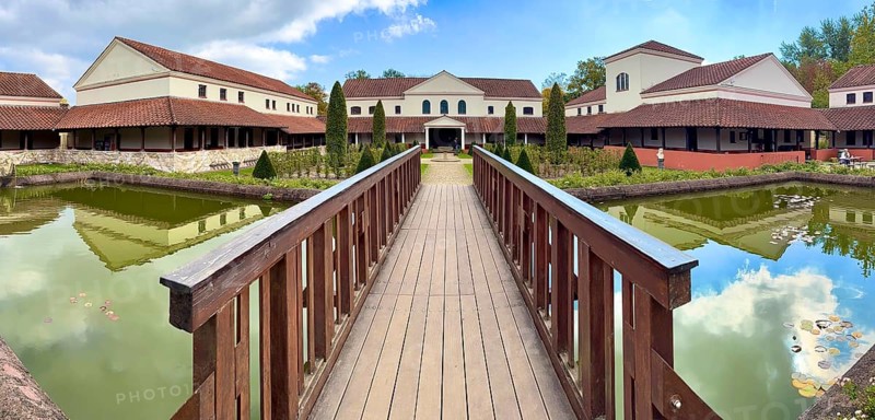 View from site of archaeological open-air museum across small wooden bridge across ornamental pond to reconstructed main building Manor House of Archaeological Park Römische Villa Borg, Perl, Saarland, Germany