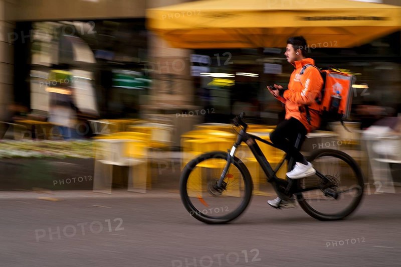 Lieferando, logo, delivery service, courier service, bicycle courier, courier driver rides with thermal backpack, e-bike, mobile phone, smartphone, movement effect, speed, blurred, Stuttgart, Baden-Württemberg, Germany