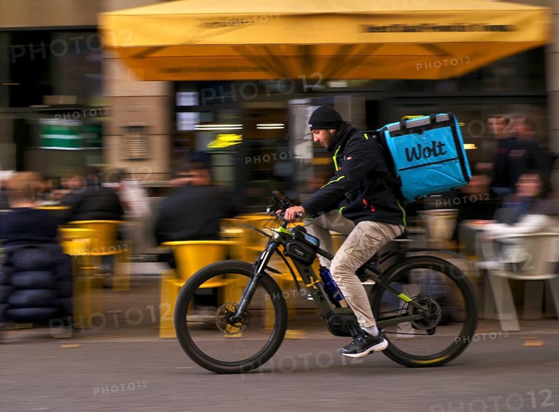 Wolt Enterprises Oy, logo, delivery service, courier service, bicycle courier, courier driver rides with thermal backpack, e-bike, mobile phone, smartphone, movement effect, speed, blurred, Stuttgart, Baden-Württemberg, Germany