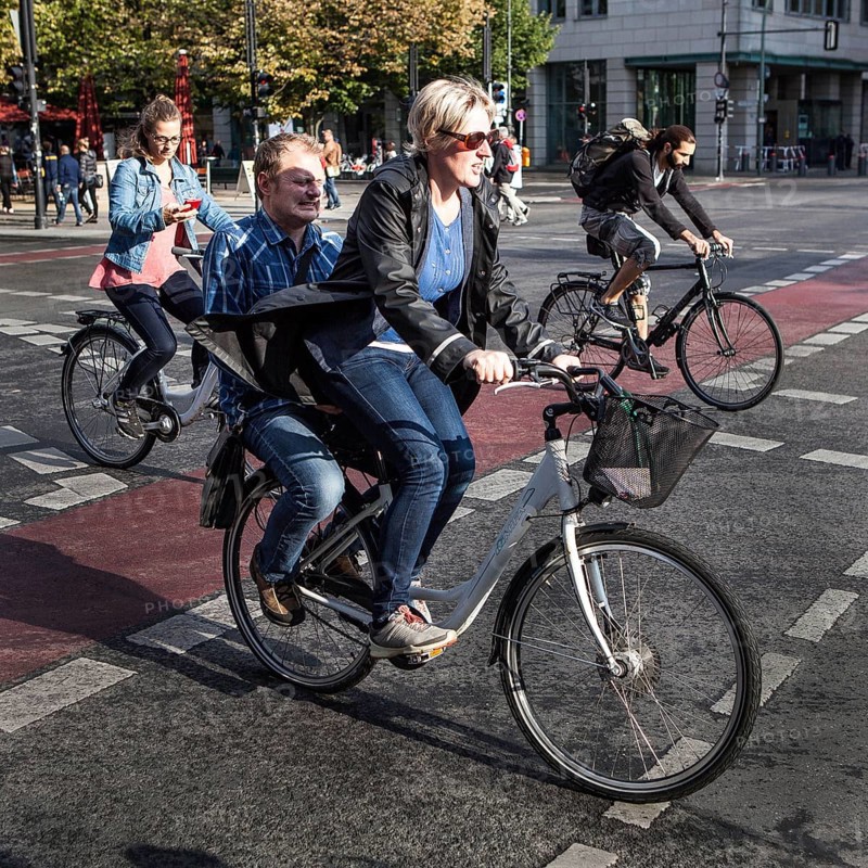 DEU Germany Germany Berlin cyclist transports a man on the luggage rack while another woman watches her smartphone. Everyone is driving on the road 'Unter den Linden' towards Brandenburg Gate