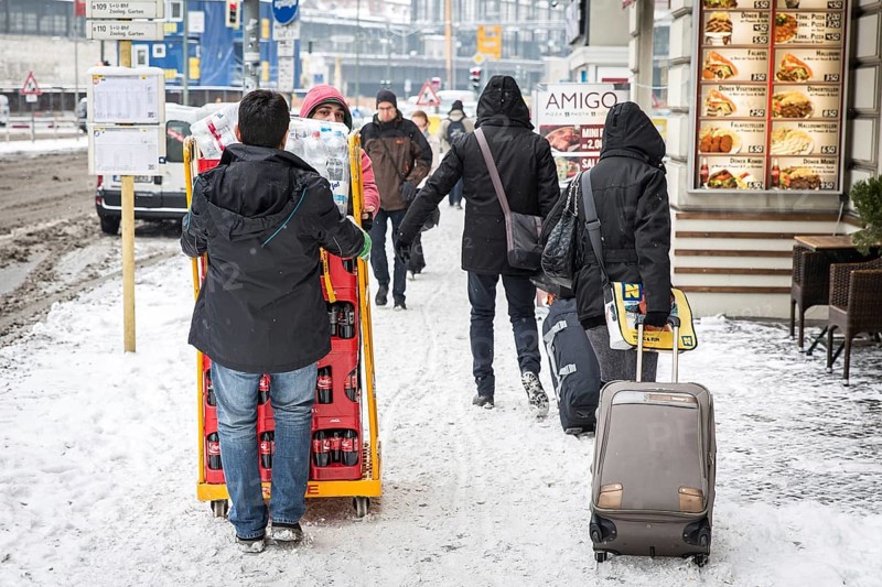 DEU Germany Germany Berlin Travelers with wheeled suitcases pass by beverage purveyors on a snow-covered sidewalk near Zoo Station