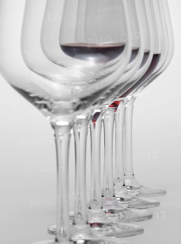Some wine glasses standing consecutive in a row in light grey back, one partly filled with red wine