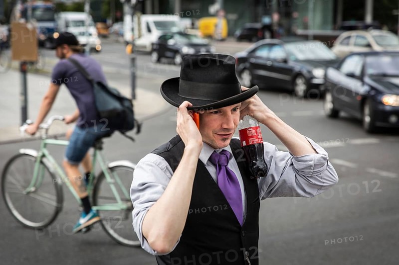 DEU Germany Germany Berlin A man protects his hat from a gust of wind while talking on the phone and holding a coke bottle