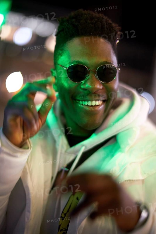 Greenlit African man wearing sunglasses at night as shade of anonymity, approved immigration, integration, laughing, happy, giggle, funny, joyful, optimistic, anticipation, expecting, expectation, optimism, proactive, can do attitude, outgoing, charming, big smile, open-minded, optimist, immigrant, foreigner, approval, green, environmentalism, environmental attitude, sustainability, green as color for sustainable environment, candid happiness, jolly, excited, excitement, high spirited, elated, motivated, motivation, good mood, movement, cheerful, cheerfulness
