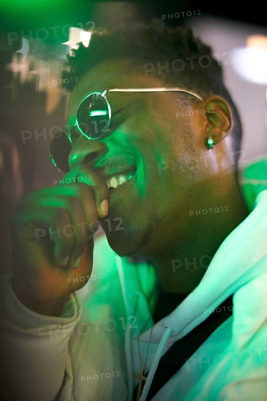 Greenlit African man wearing sunglasses at night as shade of anonymity, laughing, happy, giggle, funny, joyful, optimistic, optimist, approved immigration, integration, immigrant, foreigner, approval, green, environmentalism, environmental attitude, sustainability, green as color for sustainable environment, amusement, amused