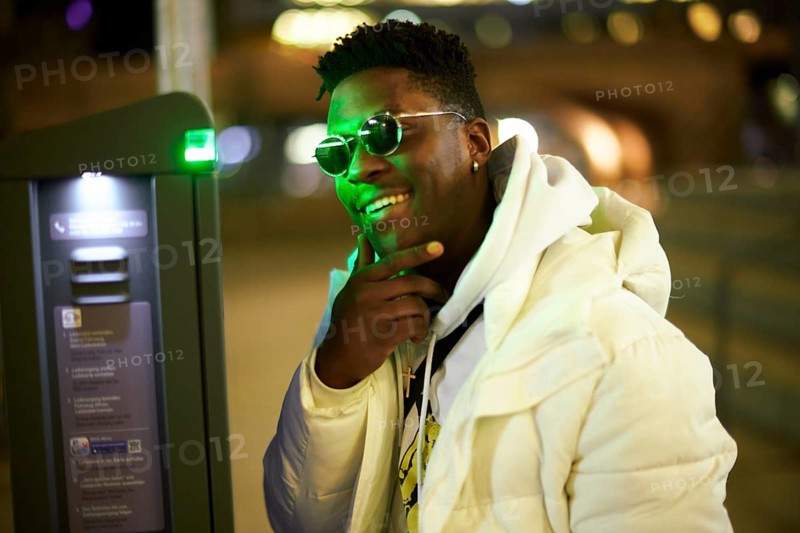 Thoughtful greenlit African man at parking ticket machine pausing as symbol for approved immigration and ability to pause and recharge, wearing sunglasses as shade of anonymity, scepticism, thinking, pondering, thoughts, scpetic, sceptical, hesitation, hesisting, hand on chin, integration, smile, smiling, amused, good mood, optimist, optimistic, optimism