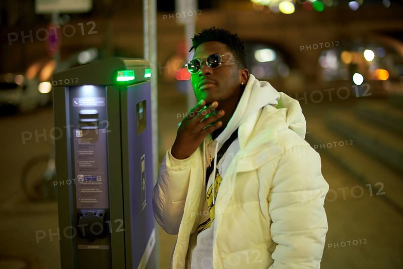 Thoughtful greenlit African man at parking ticket machine pausing as symbol for approved immigration and ability to pause and recharge, immigrant, wearing sunglasses as shade of anonymity, scepticism, thinking, pondering, thoughts, scpetic, sceptical, hesitation, hesisting, hand on chin, approval, integration, green as color for sustainability and environmentalism, sustainable mobility transition