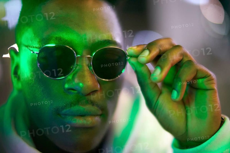 Greenlit African man wearing sunglasses at night as shade of anonymity, coolness, approved immigration, integration, immigrant, foreigner, approval, green, environmentalism, environmental attitude, sustainability, poised, serious, calm, close-up, green as color for sustainable environment and optimism and being greenlit approved