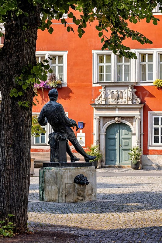 A statue in a square in front of a red, historic building with green door and blue windows, Johann Sebastian Bach memorial in the old town of Arnstadt in Thuringia