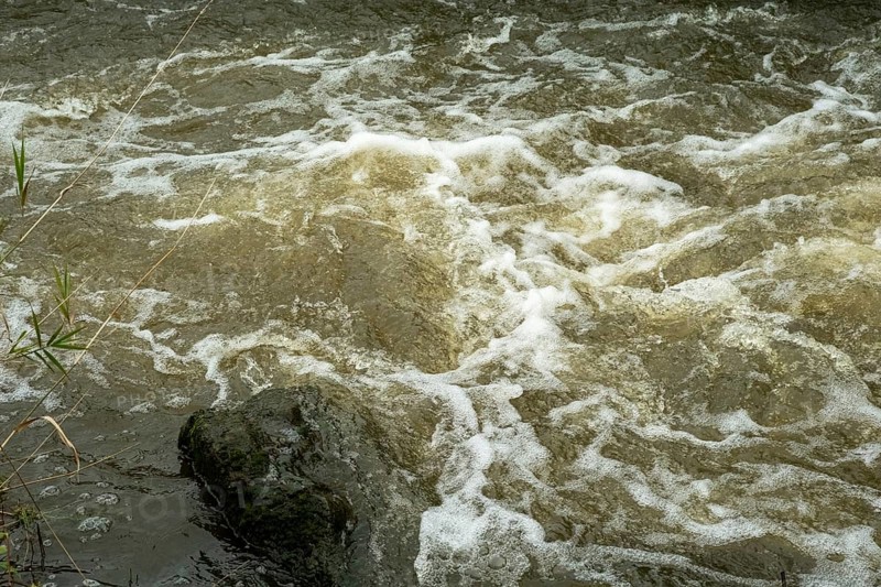 Borken-Hoxfeld, NRW, Germany, The scene features a fast-flowing river with clear water splashing over smooth rocks. Bubbles form as the water rushes past, surrounded by grassy edges. Light reflects off the surface