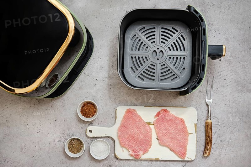Freshly prepared pork loin cutlets sit on a cutting board beside spices. An air fryer awaits on the kitchen counter to transform the meat into a delicious meal