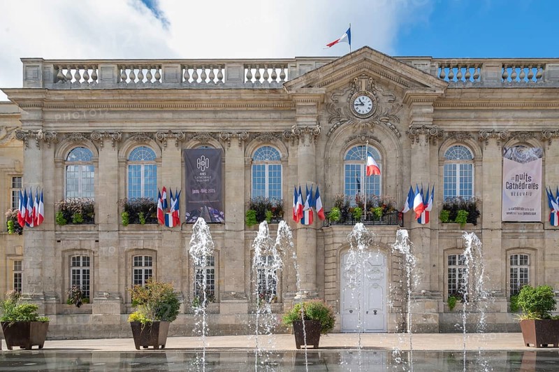 The classicist façade of Beauvais Town Hall (Hotel de Ville) dominates Place Jeanne Hachette. Floor fountains can be seen in action in the foreground. The building is decorated with numerous tricolor flags (drapeaux francais), under a cloudy blue sky in Beauvais, Oise department (60), Hauts-de-France region, France