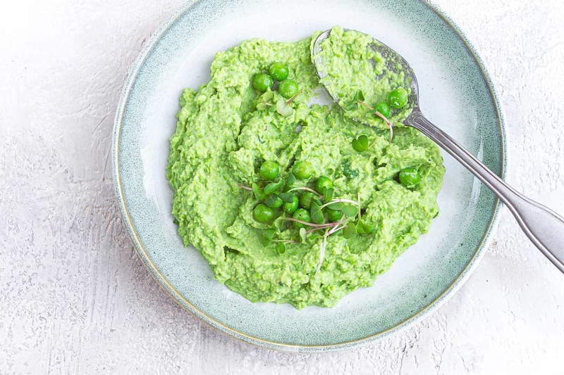 A vibrant green pea puree fills a shallow bowl, topped with fresh peas and microgreens. The dish looks fresh and inviting, ideal for a light, nutritious meal at any time