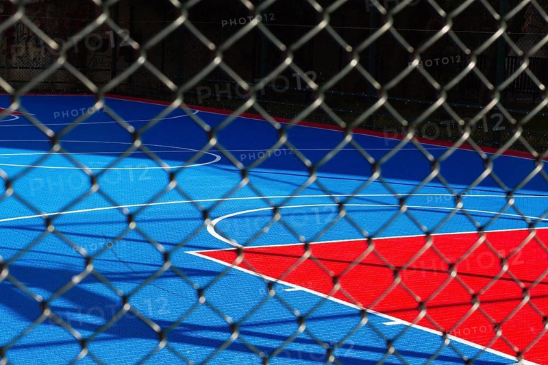 Brightly colored basketball court features blue and red sections, seen through a chain-link fence. Sunlight casts shadows on the court, indicating a sunny day in the city