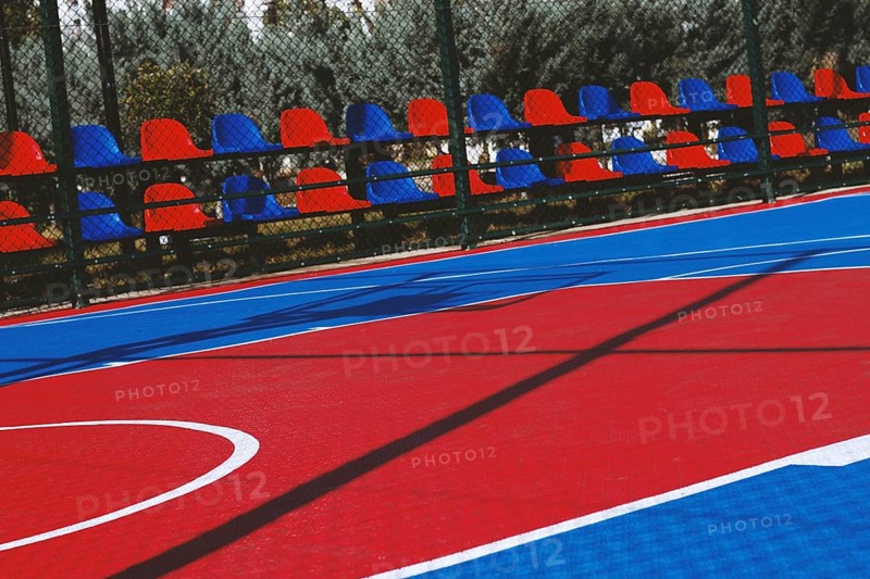 A brightly colored basketball court features bold red and blue sections under the clear sky. Nearby, empty chairs line the sidelines, ready for spectators on a sunny day