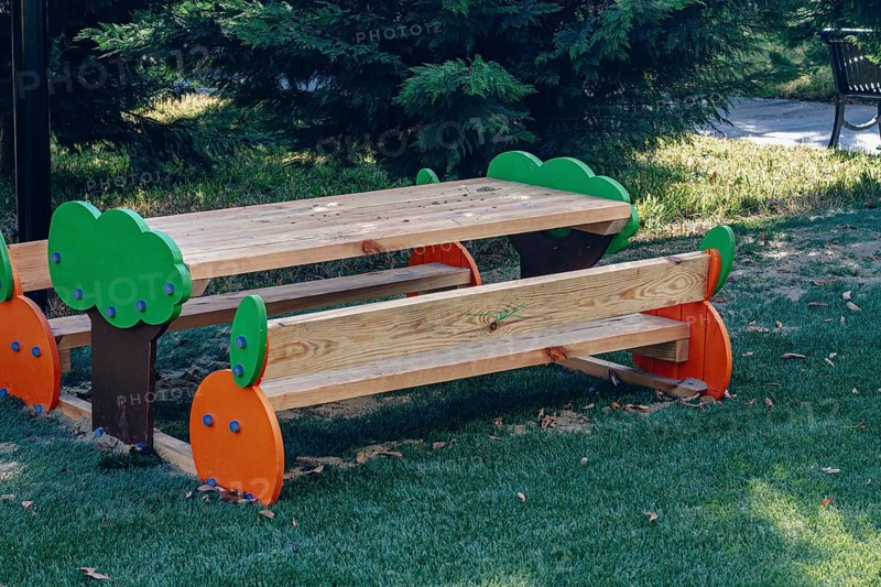 A vibrant wooden picnic table with colorful tree designs stands in a green park, surrounded by grass and trees under clear blue skies on a sunny day