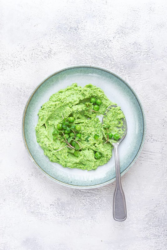 A vibrant green pea puree fills a shallow bowl, topped with fresh peas and microgreens. The dish looks fresh and inviting, ideal for a light, nutritious meal at any time