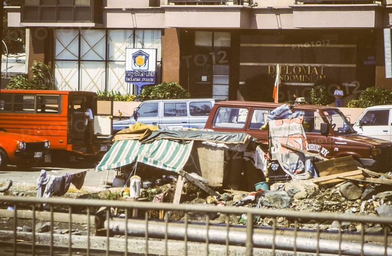 Homeless people have built an emergency shelter consisting of a wooden box and awning on the middle strip of a multi-lane road, behind it luxury apartments, Manila in 1995, Philippines