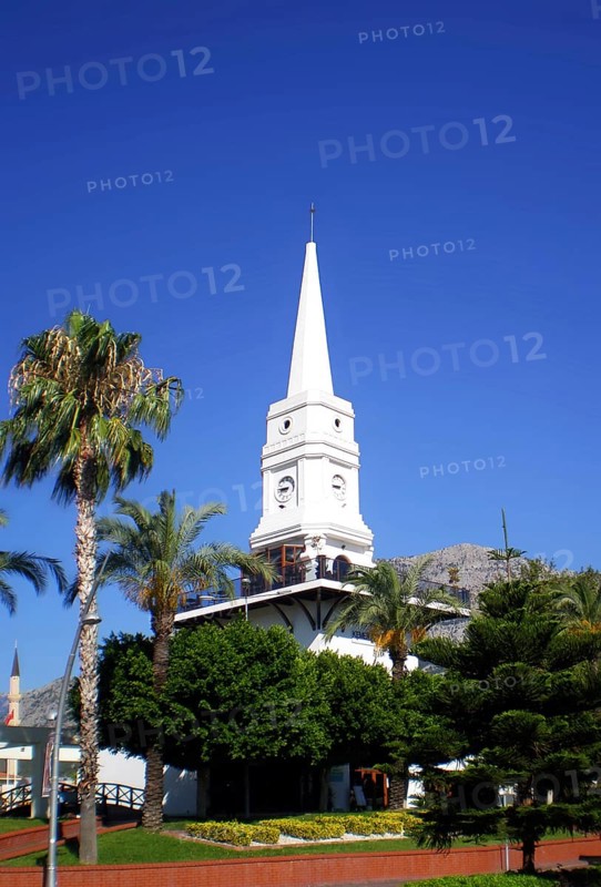 A tall white clock tower surrounded by palm trees, one of the main architectural landmarks for tourists and travelers of the resort town of Kemer (Antalya Province) and the entire eastern Mediterranean Turkish Riviera. Kemer, Turkey