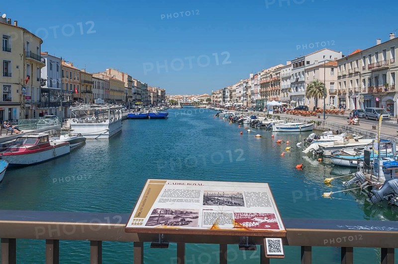 The Canal de Sete, formerly Canal Royal, stretches between the historic house facades, with an information board on the history of the canal in the foreground and numerous moored boats on the banks, seen from the Pont de la Savonnerie under a blue sky in Sete, Herault department, Occitanie region, France