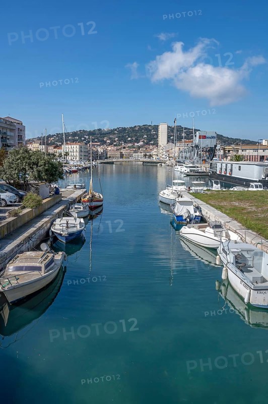 View from Pont des Dockers across the Canal de la Peyrade with moored boats along the quays towards the skyline of the city center with the wooded Mont Saint Clair in the background under a blue sky in Sete, Herault department, Occitanie region, France