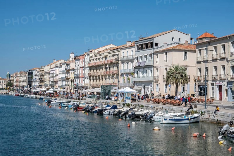 The lively promenade of Quai Leopold Suquet along the Canal de Sete, formerly Canal Royal, with a series of historic house facades, restaurants with outdoor restaurants and numerous moored boats on the shore under a blue sky in Sete, Herault department, Occitanie region, France