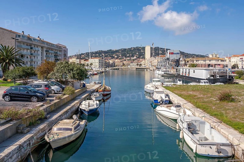 View from Pont des Dockers across the Canal de la Peyrade with moored boats along the quays towards the skyline of the city center with the wooded Mont Saint Clair in the background under a blue sky in Sete, Herault department, Occitanie region, France