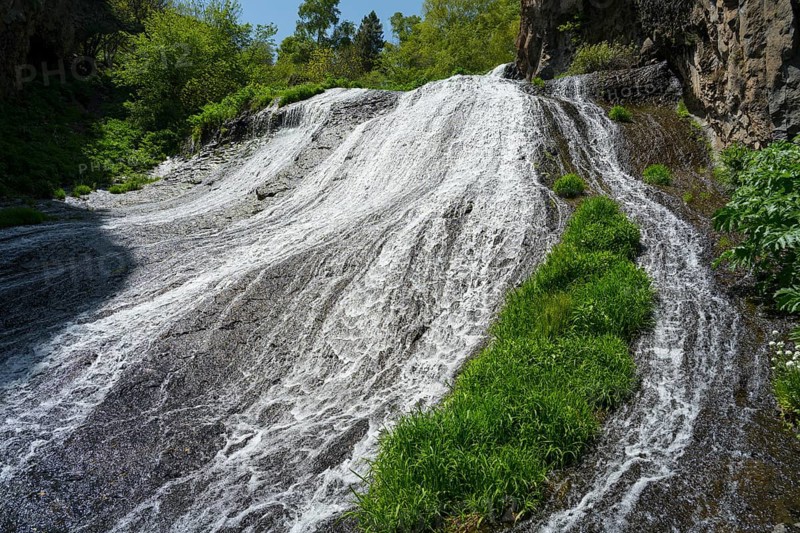 A wide waterfall flows steeply down, flanked by green plants and rocks in nature, Jermuk Waterfall, Vayots Dzor Province, Armenia