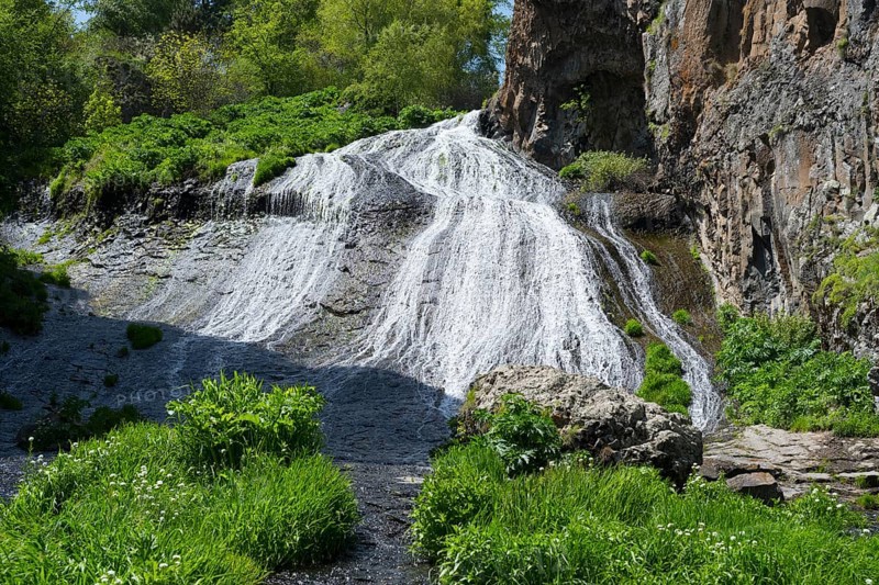 A bright waterfall snakes through green plants and rocks in sunny nature, Jermuk Waterfall, Vayots Dzor Province, Armenia