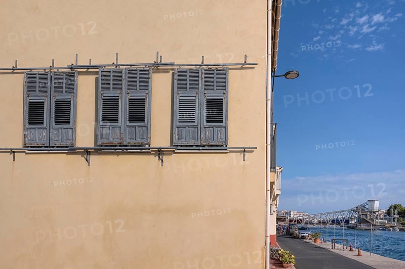Yellow house façade with three gray wooden shutters to move and a view across the Canal de Sete (formerly Canal Royal) to the distinctive steel turnstile Pont Marechal Foch and the port area in La Pointe Courte in Sete, Herault department, Occitanie region, France