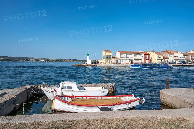 Two moored fishing boats in the foreground with a view across the Canal de Sete, formerly Canal Royal, of the colorful houses and the white-green lighthouse of La Pointe Courte, while a blue sightseeing boat passes by, under a blue sky in Sete, Herault department, Occitanie region, France