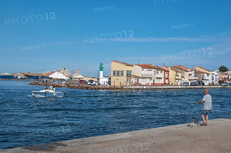 An angler looks from the banks of the Canal de Sete, formerly Canal Royal, across to the green-white lighthouse of La Pointe Courte, while a man steers his motorboat out onto the Etang de Thau, Sete, Herault department, Occitanie region, France