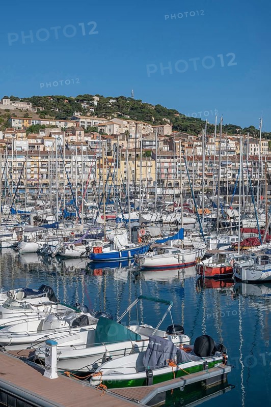 Densely packed sailboats and yachts with numerous masts in the port of Sete, with the densely built old town and Mont Saint-Clair in the background under bright blue skies in Sete, Herault department, Occitanie region, France