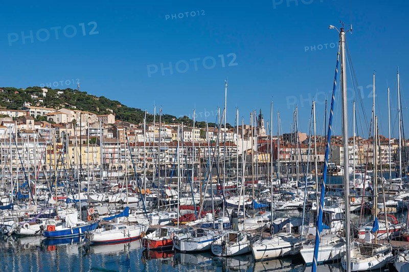 Densely packed sailboats and yachts with numerous masts in the port of Sete, with the densely built old town and Mont Saint-Clair in the background under bright blue skies in Sete, Herault department, Occitanie region, France