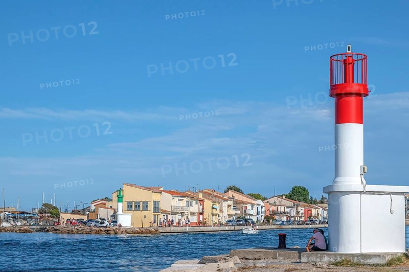Men sitting at the red and white lighthouse at the port entrance of the Canal de Sete, formerly Canal Royal, with the typical, colorful fishermen's houses of the La Pointe Courte quarter under a blue sky in Sete, Herault department, Occitanie region, France