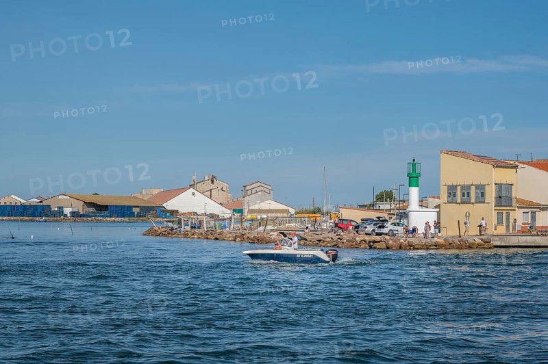 A motorboat with people on board steering it sails in front of the white-green little lighthouse and a yellow fisherman's house at the port entrance of the Canal de Sete, formerly Canal Royal, at La Pointe Courte with industrial buildings in the background under a blue sky in Sete, Herault department, Occitanie region, France