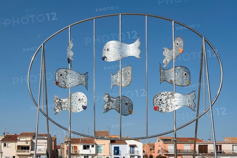 The modern metal sculpture Les Daurades Miroirs by Dominique Dore on the Plage de la Lagette shows silver mirror fish in an oval frame, with the colorful houses of the La Pointe Courte district in Sete, Herault department, Occitanie region, France