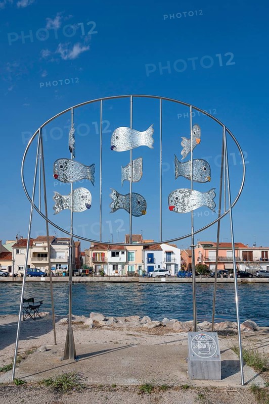 The modern metal sculpture Les Daurades Miroirs by Dominique Dore on the Plage de la Lagette shows silver mirror fish in an oval frame, with the colorful houses of the La Pointe Courte district in Sete, Herault department, Occitanie region, France