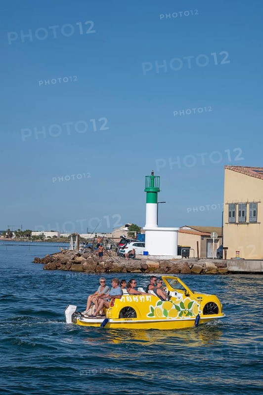 Sightseeing on the water: a yellow water car with tourists and lemon decorations rides on the Canal de Sete, formerly Canal Royal, in front of a white-green lighthouse and fishermen's houses in La Pointe Courte under a bright blue sky in Sete, Herault department, Occitanie region, France
