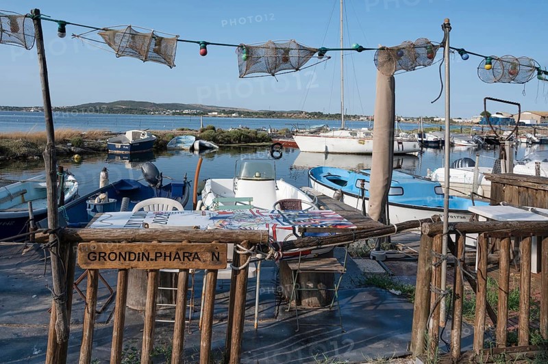 A fisherman's sitting area for an aperitif with tables and chairs as well as fishing nets as decoration, framed by fishing boats in the small port of La Pointe Courte with a view of the Etang de Thau under a blue sky in Sete, Herault department, Occitanie region, France
