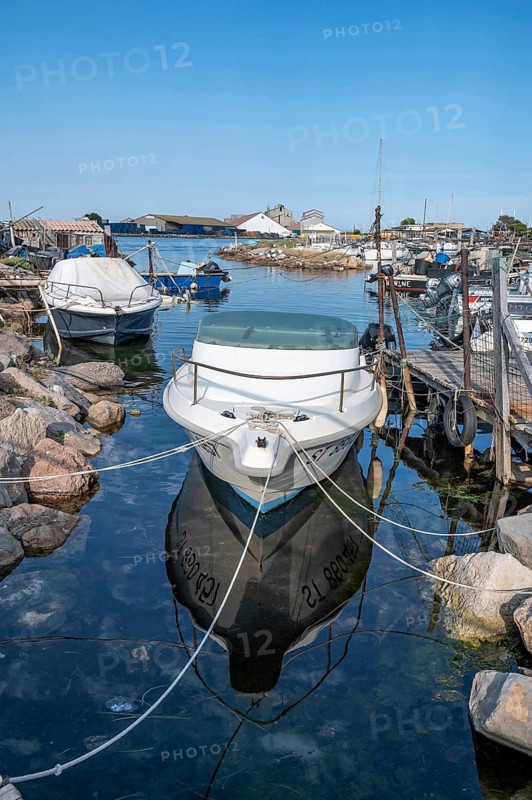 A white moored motor boat with reflection in the calm water of a small harbor of the traditional fishing port of La Pointe Courte between rocks and wooden walkways, with other fishing boats and industrial buildings under a blue sky in Sete, Herault department, Occitanie region, France