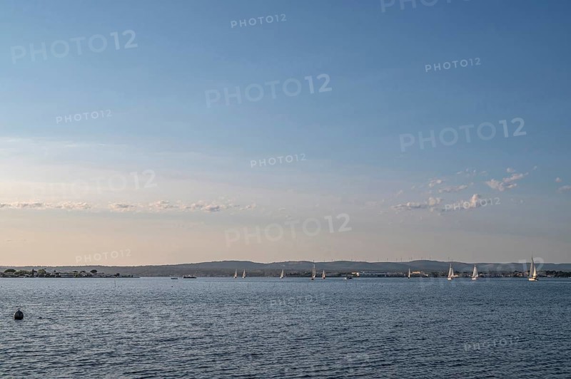 Wide-angle landscape view of Etang de Thau with several sailboats on the water sailing towards the skyline of the Balaruc coast, seen from the banks of the Le Barrou district under blue summer skies in Sete, Herault department, Occitanie region, France