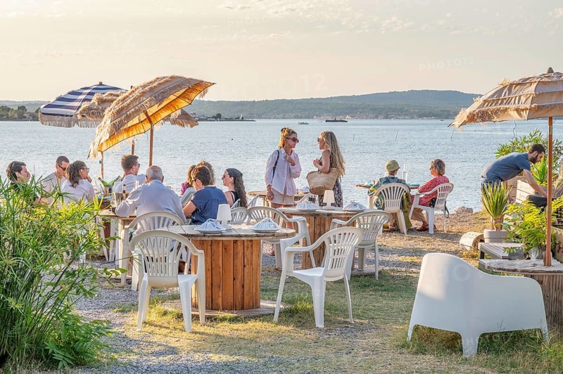 Summer scene on the terrace of the Guinguette A la bonne Franquette with guests sitting at tables under umbrellas and talking, with a view over the Etang de Thau in golden evening sun, Le Barrou district, Sete, Herault department, Occitanie region, France
