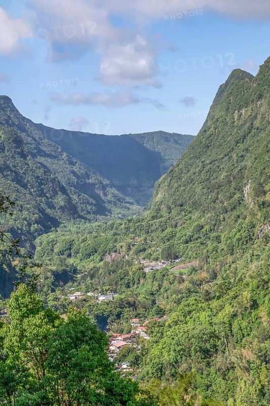 View from Department 52 of the massive, steep Remparts rock faces of Cirque du Salazie and a hamlet at the bottom of the valley on a sunny valley with some clouds in the blue sky, Cirque de Salazie, Tropical Highlands, La Reunion, France