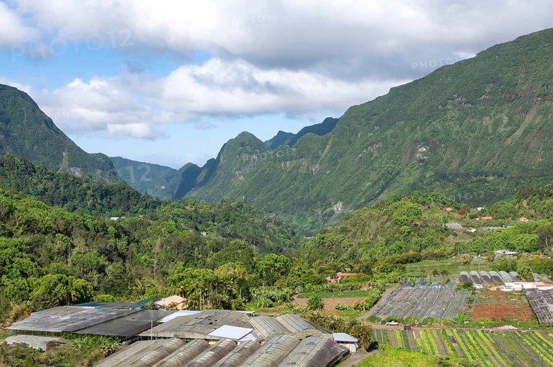 Tropical valley in Cirque de Salazie with intense green Rempart cliffs and slopes surrounded by low-hanging clouds under a blue sky, while agriculture, wax houses and houses shape the low-lying valley floor in the foreground, Cirque de Salazie, Tropical Highlands, La Reunion, France