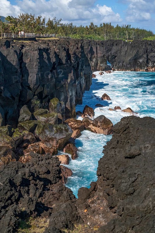 Massive, black lava cliffs rise steeply out of the Indian Ocean at Cap Mechant, where white surf washes around the volcanic rock, while in the background a series of palm trees and tropical trees separate the lava coast from the land side, Cap Mechant, South Coast, La Reunion, France