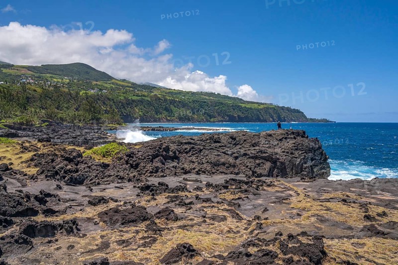 An angler stands on a massive rock of the black lava coast and looks out at the roaring Indian Ocean as the surf white washes around the volcanic rock and in the background the steep, green cliffs near the Marine de Vincendo rise into the sky, Marine de Vincendo, South Coast, La Reunion, France