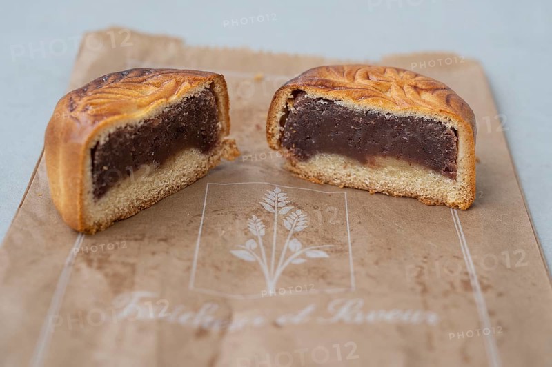 Close-up of a mooncake (mooncake) cut in the middle with a thick layer of dark red bean paste and a light shortcrust pastry base, presented on brown recycled paper with stylized print (Saveurs et Bonheur) as a symbol of Chinese culinary culture on the island, Saint-Pierre, La Reunion, France
