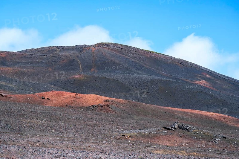 The Route du Volcan leads through the vast, black volcanic desert of the Plaine des Sables, where a black-brown lava hill rises with a path in the flank, whose red and orange Scoria deposits in the foreground shape the geological lunar landscape, Plaine des Sables, Highlands, La Reunion, France